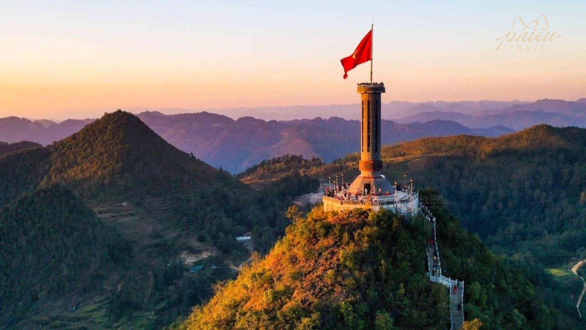 A stunning panoramic view of the Lung Cu Flag Tower in Ha Giang, featuring the large Vietnamese flag atop the stone tower on Dragon Mountain at sunset