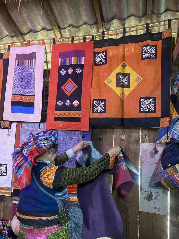 A local ethnic minority woman in colorful traditional attire examines a handwoven textile in a rustic shop, surrounded by vibrant patterned fabrics hanging on the wall and small wooden toys below.